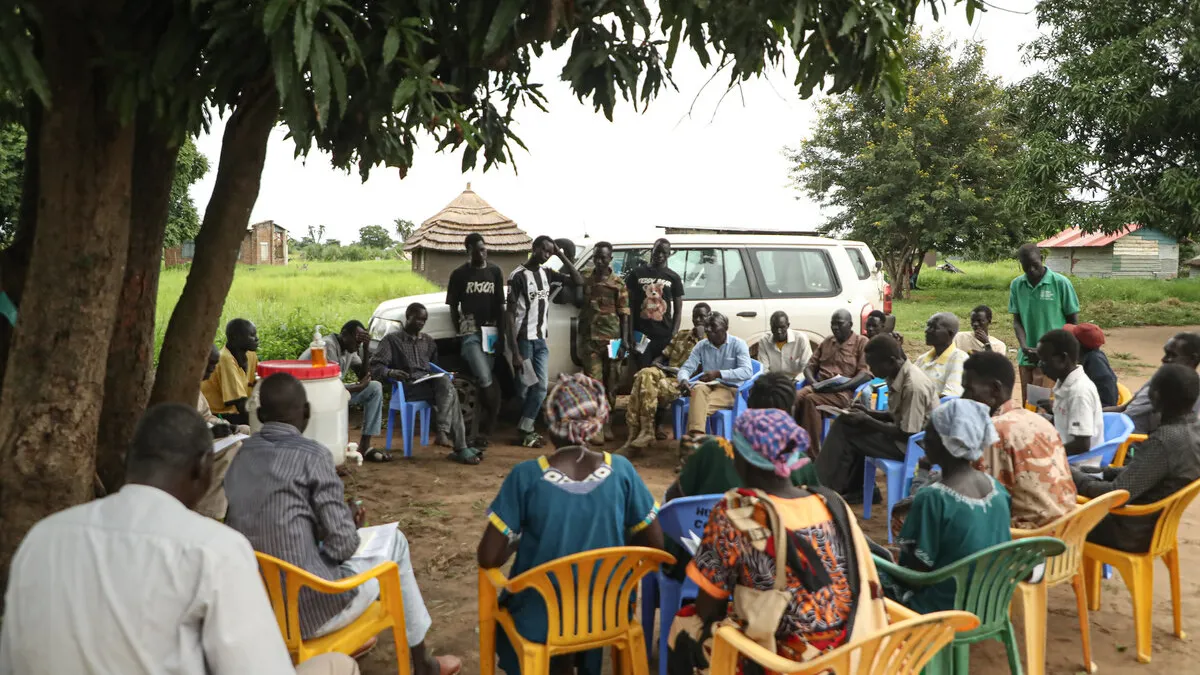 A visiting UNMISS team engaged with local communities in Gondokoro, where land conflicts between farmers and cattle herders from elsewhere have led to tensions and insecurity. Community meeting held outdoors under a tree, with people seated in a circle on plastic chairs and several individuals standing near a white vehicle in the background.