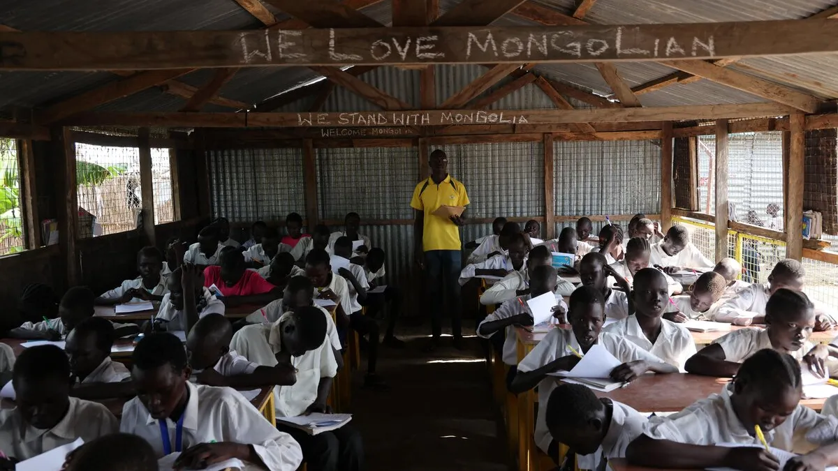 After 13 years of protecting civilians in Bentiu, the relationship between Mongolian peacekeepers and local communities is so strong that the children can even speak the Mongolian language. A classroom with students in white uniforms seated at wooden desks, writing in notebooks, while an instructor stands at the front under a corrugated metal roof with chalk writing above.
