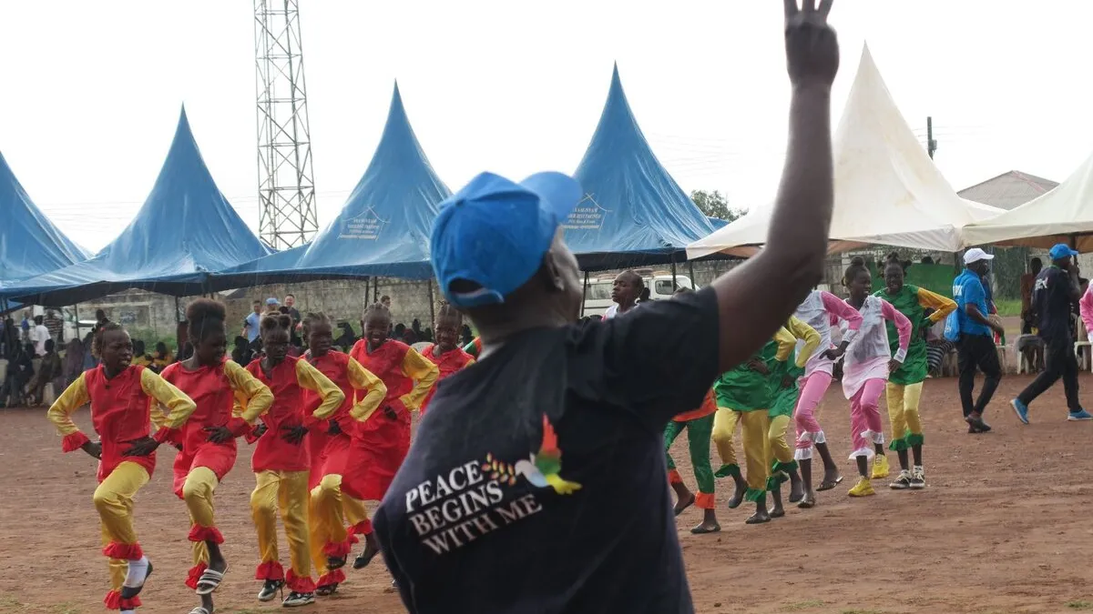 Wau’s third annual marathon, gathering over 2,000 participants, launched the UNMISS campaign "Peace Begins With Me” in Western Bahr El Ghazal. Outdoor event with a person wearing a shirt that reads “Peace Begins With Me” raising a hand, while groups of performers in colorful outfits dance in formation near blue tents.