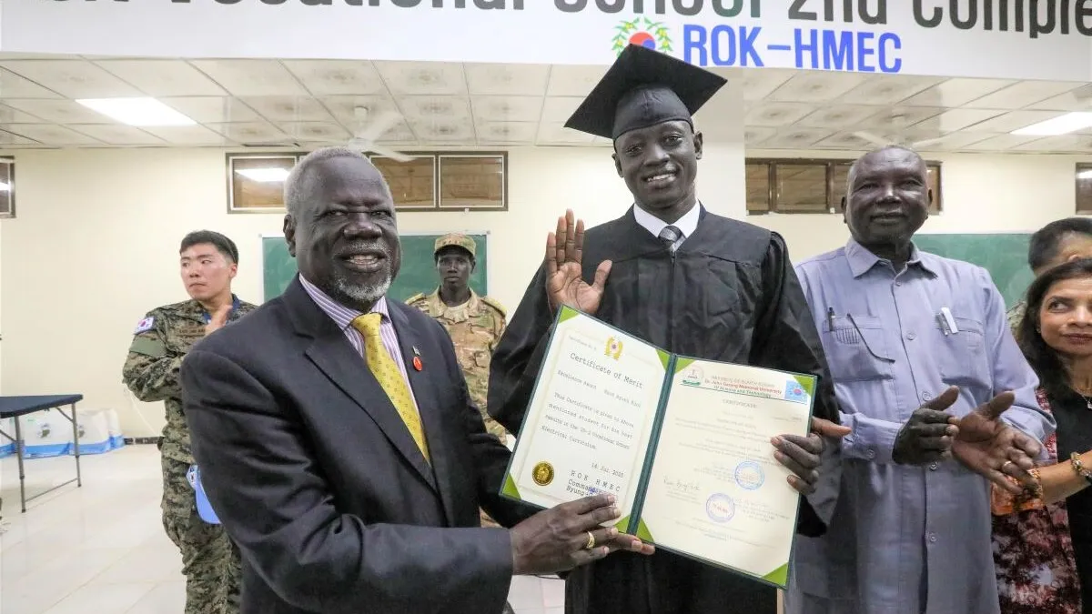 Youth in South Sudan often have difficulties finding learning opportunities and jobs. In Bor, peacekeepers from South Korea have conducted (Bor) 3-month-long vocational trainings for young, and now optimistic, women and men. In Akobo, UNMISS has funded a similar initiative. Graduation ceremony indoors with a person in a cap and gown holding a certificate, surrounded by others, with a banner for a vocational school visible above.