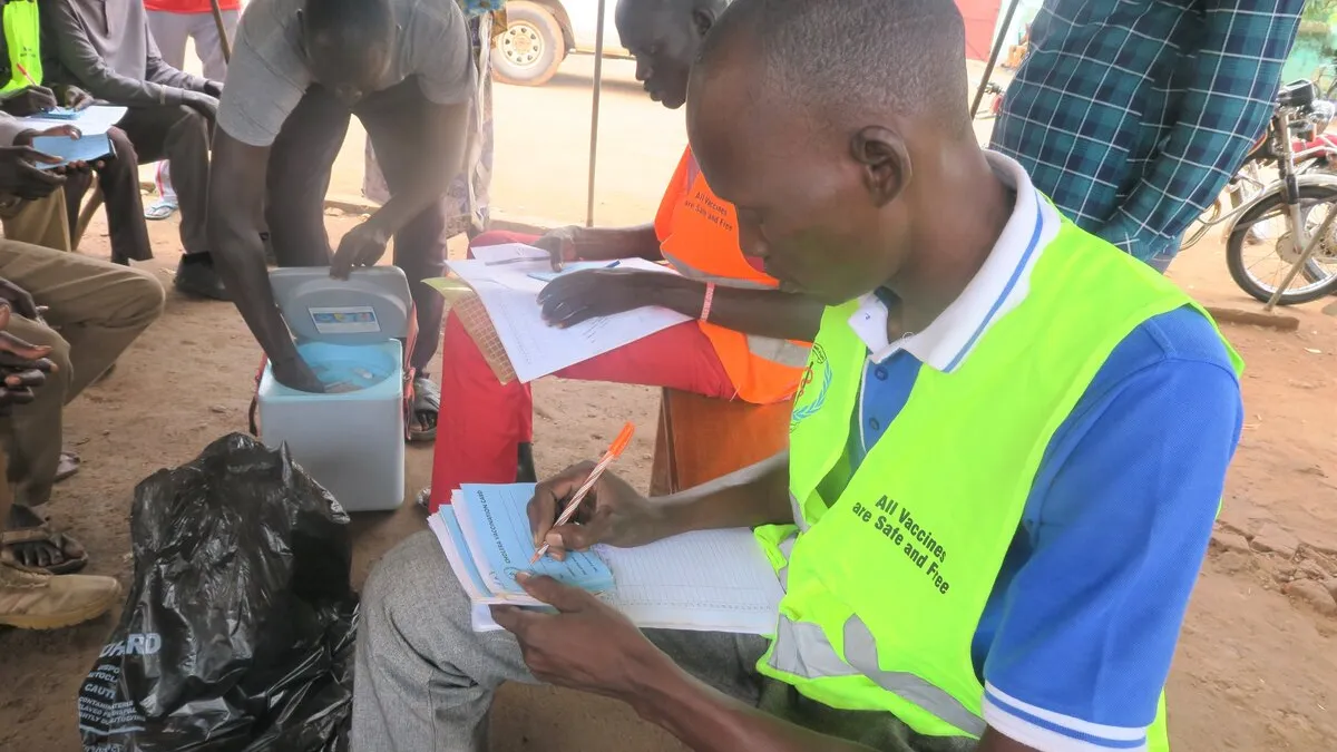 With a capacity of 150 inmates, Aweil Central Prison in Northern Bahr El Ghazal currently houses over 700 prisoners in crowded, unhygienic conditions. With cholera running rampant in the region, UNMISS advocated with local authorities and the World Health Organization to vaccinate all prisoners and their guards. Health worker wearing a bright vest writes in a register during a vaccination campaign, with a cooler box and other people seated nearby.