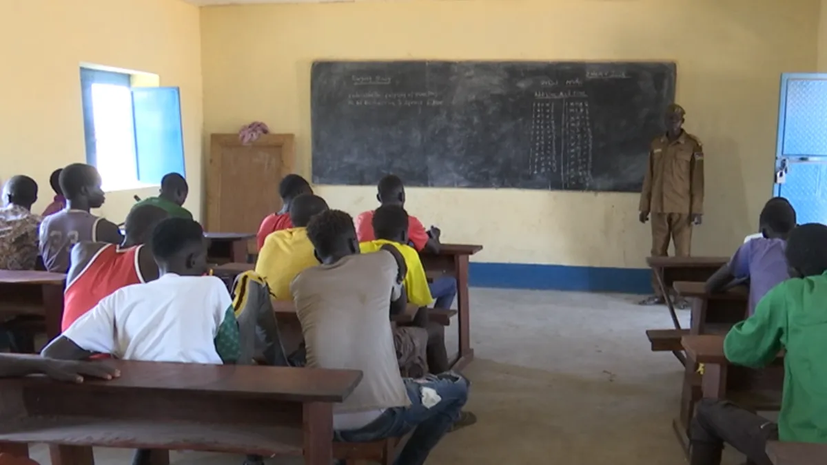 At the prison in Kuajok, the vocational trainings initiated by UNMISS previously have an impact by offering inmates to develop skills in tailoring, farming and accounting. Classroom scene with students seated at wooden desks facing a chalkboard, where mathematical calculations and notes are written. An instructor stands near the board in front of the class.