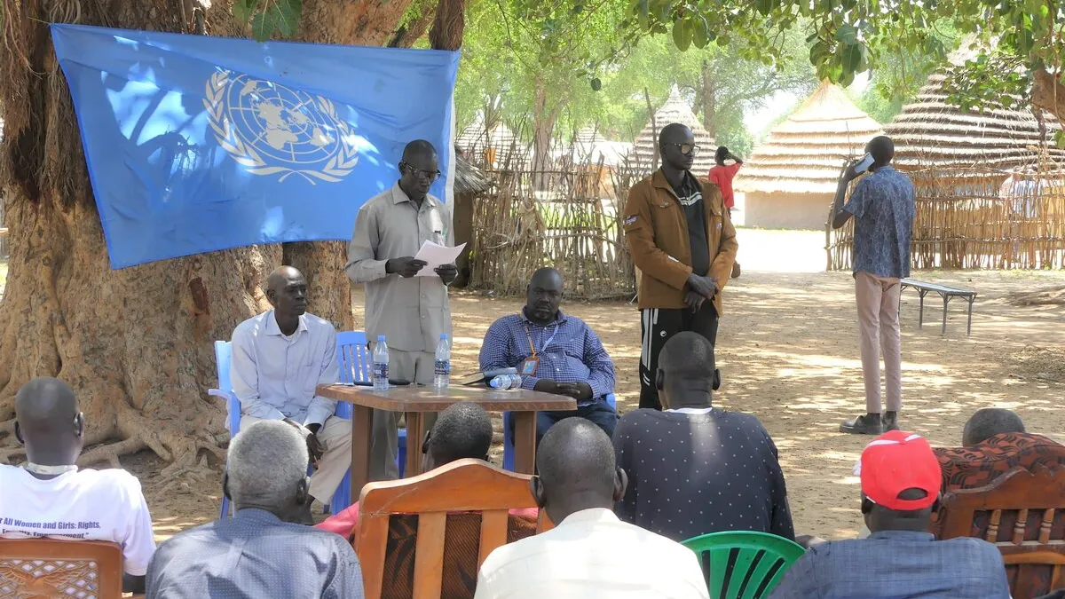 Youth leaders and other civil society activists Twic County have come together to spread the word of what previously feuding Twic and Ngok Dinka youth agreed on during a recent peace dialogue. Outdoor meeting under a large tree with a UN flag displayed. Several people are seated on chairs facing a small table where others are standing and speaking. Traditional huts are visible in the background.