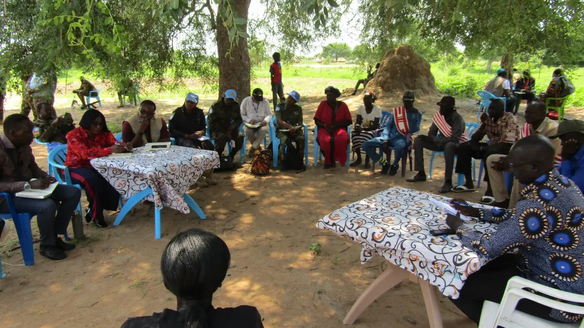 While patrolling in Panyijiar, Unity state, to help protect civilians and reduce tensions in the aftermath of air strikes, UNMISS peacekeepers hear from community members, who believe that dialogue is the only route towards a permanent peace in South Sudan. Group of people seated in a circle outdoors under a large tree, with two tables covered in patterned cloth at the center. Some participants wear blue caps, and traditional huts are visible in the background.