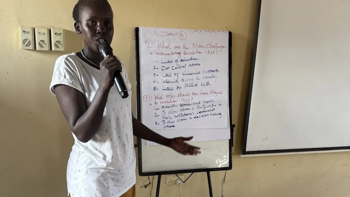 Women in Unity State have spoken out about their persistent attempts to increase and make their participation in matters related to peace and security more effective and meaningful. Person standing indoors holding a microphone and pointing toward a flip chart with handwritten notes about challenges in implementing Resolution 1325 and roles played. A blank whiteboard is positioned beside the flip chart.