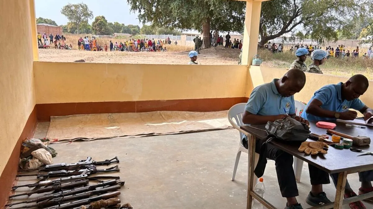 Deux hommes sont assis à une table, à côté d'une rangée d'armes à feu posées sur le sol.