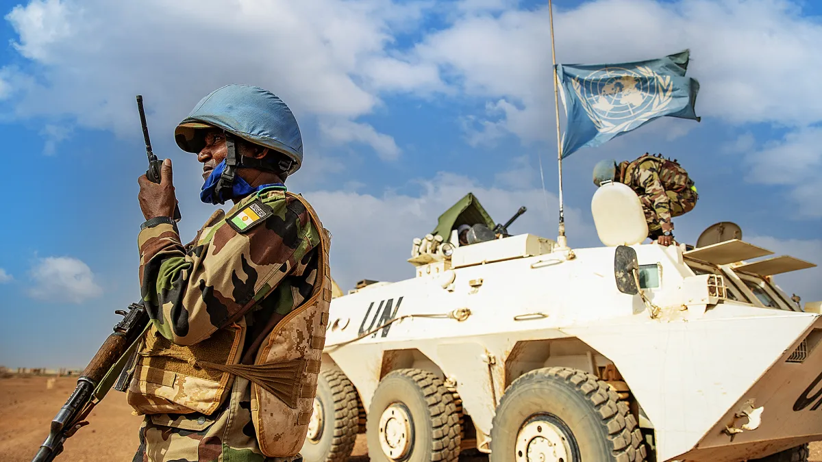A Nigerian peacekeeper in uniform and a blue helmet talking into a radio holding a gun. In the background is a white tank flying the blue UN flag. They are both in the desert with blue sky with clouds behind them.