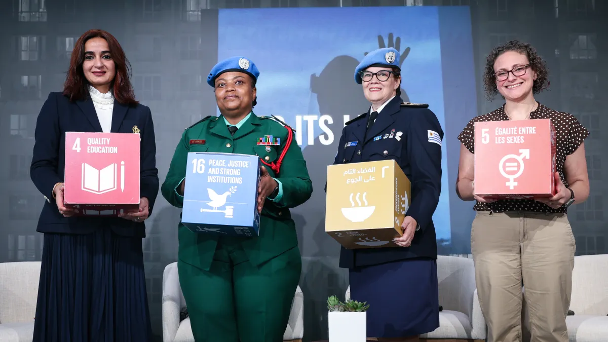 Four women peacekeepers stand side-by-side holding various SDGs
