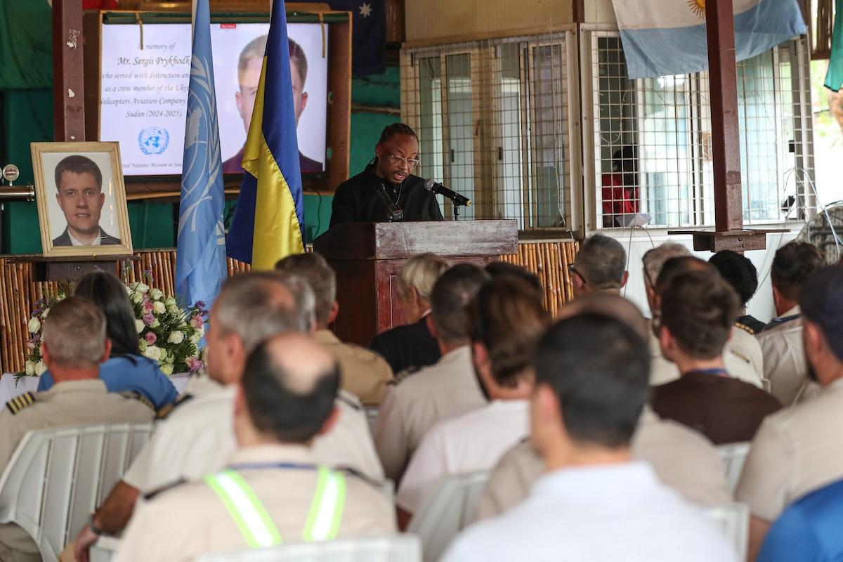 A group of people seated watching a person deliver an address at a podium in honour of a fallen peacekeeper.