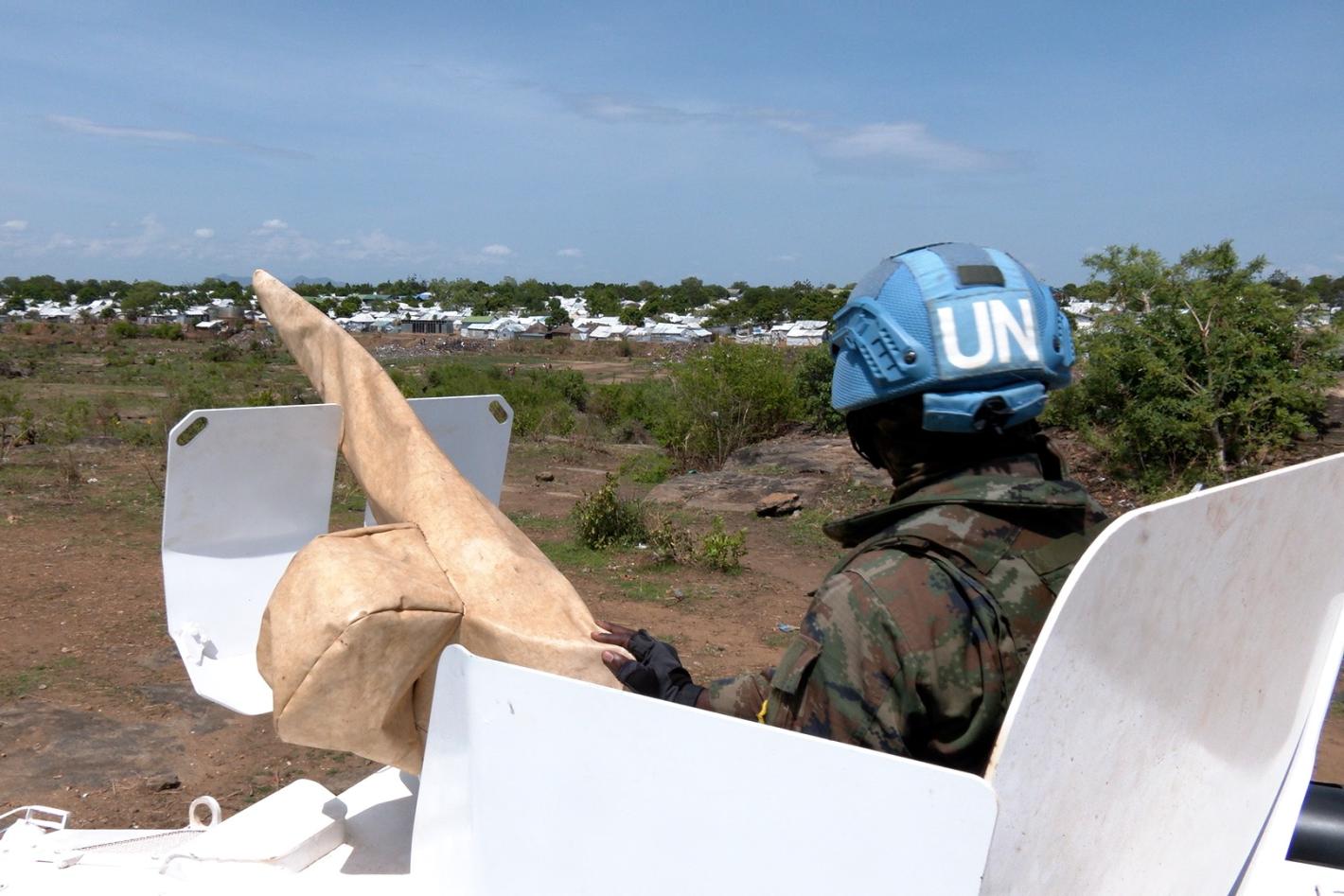 A peacekeeper peering out of the top of a tank across a grassy field. The peacekeeper is facing away from the camera.