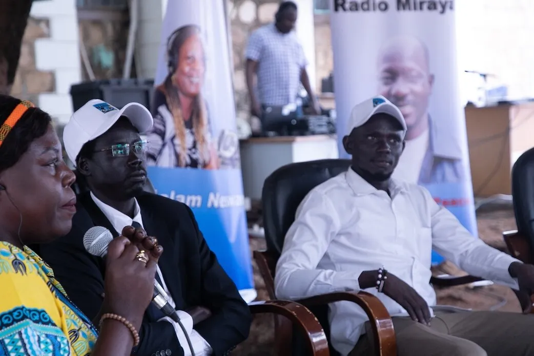 Three people seated indoors, one holding a microphone, with banners and a person standing near audio equipment in the background.