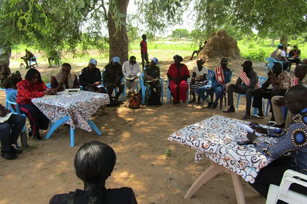 Group of people seated in a circle outdoors under a large tree, with two tables covered in patterned cloth at the center. Some participants wear blue caps, and traditional huts are visible in the background.