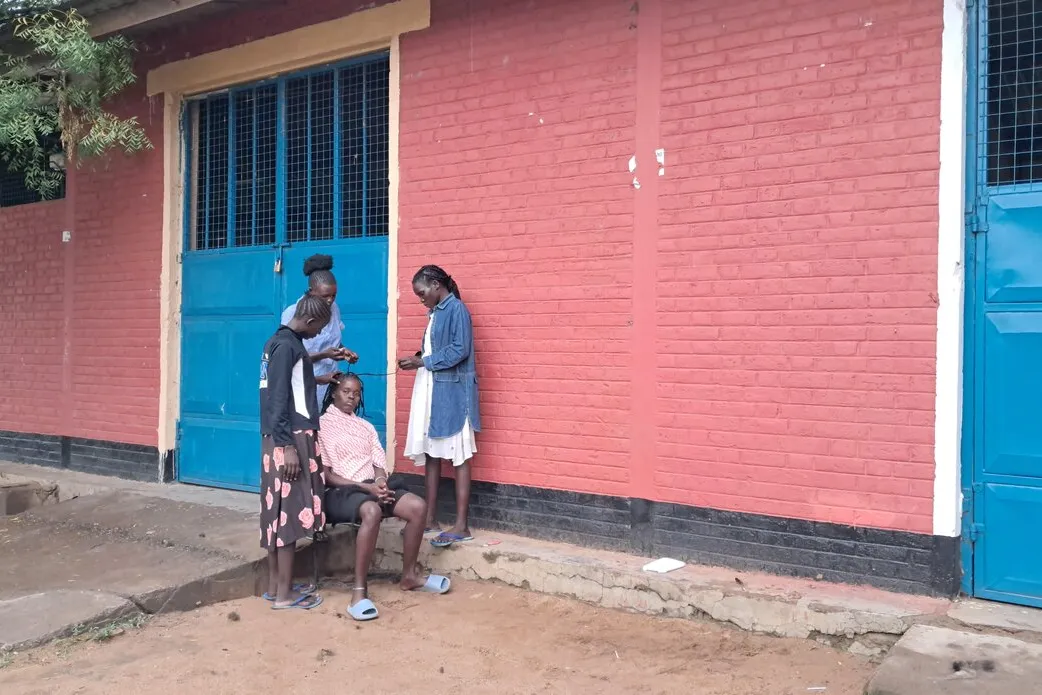 Four people stand and sit outside a building with red brick walls and bright blue metal doors, engaged in conversation near the entrance