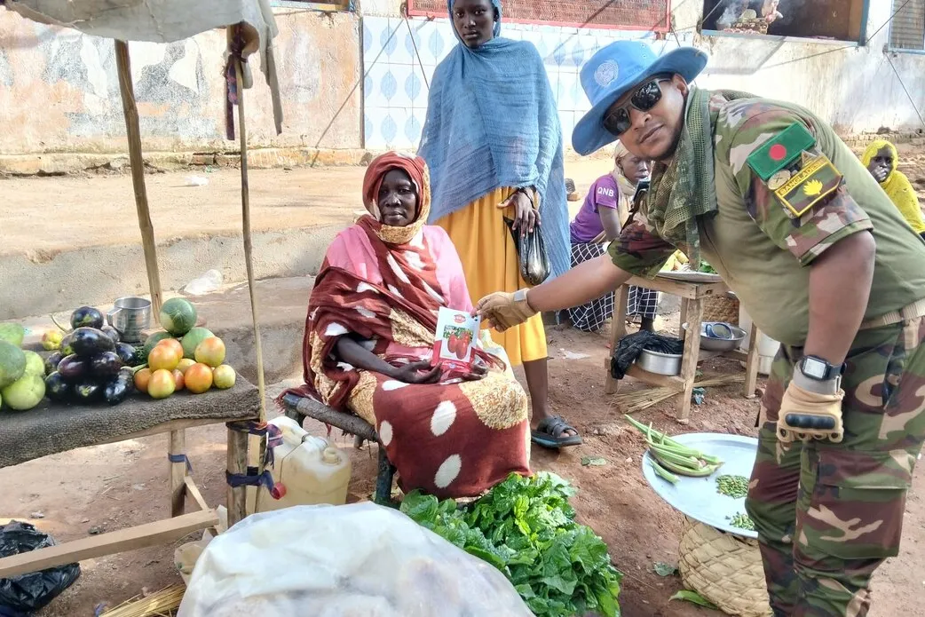 A person in camouflage uniform hands a leaflet to a seated vendor at an outdoor market stall with fruits and vegetables displayed on a mat
