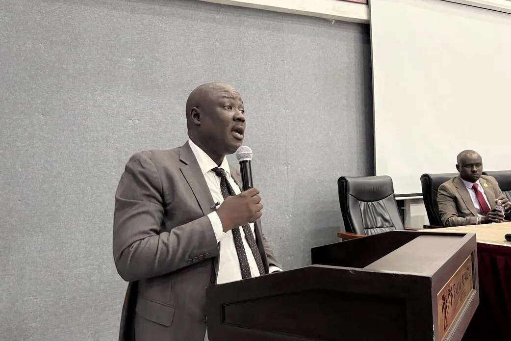 A person in a suit speaks at a podium with a microphone in a conference room, while another person sits at a table with documents and a microphone in front.