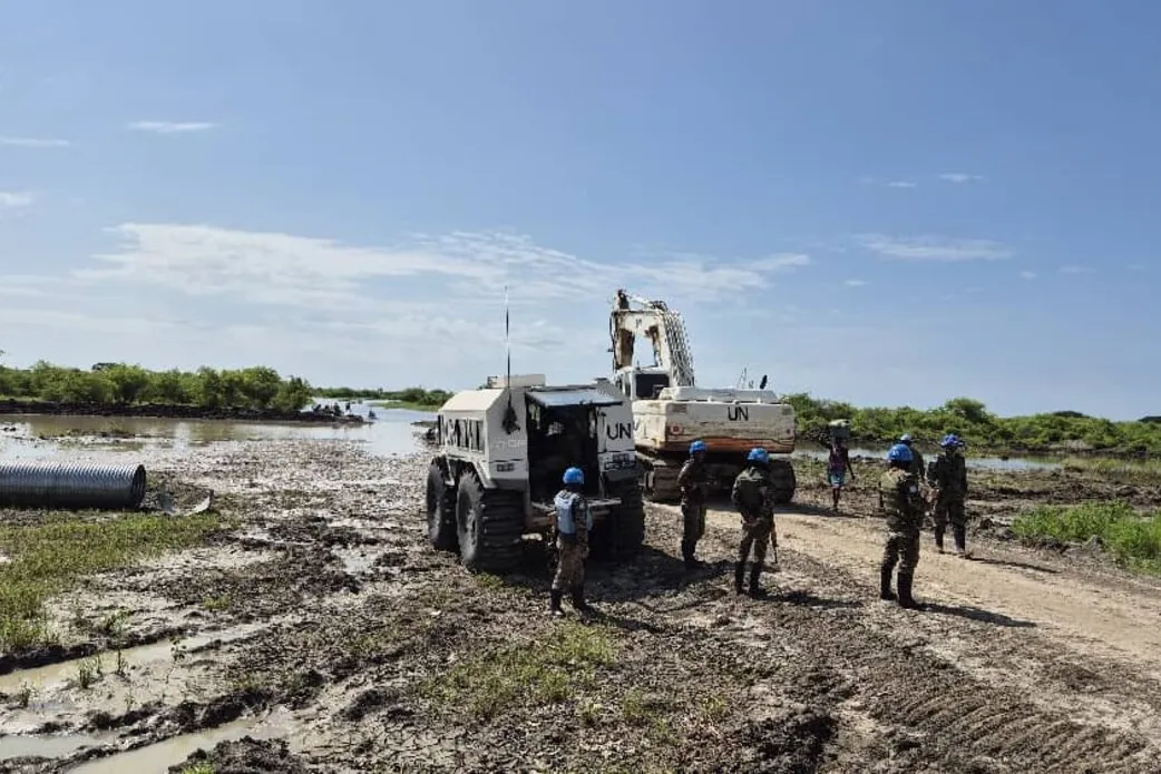 UN peacekeepers in blue helmets stand on a muddy road near heavy UN machinery and pipes, with water and greenery in the background.