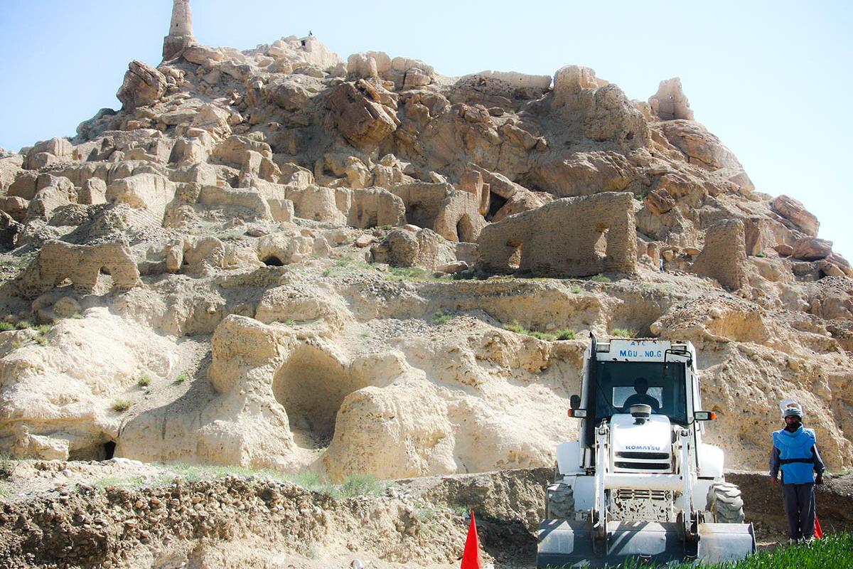 A rocky hillside with ancient-looking stone ruins carved into the cliff face, while a white construction vehicle and a worker stand at the base