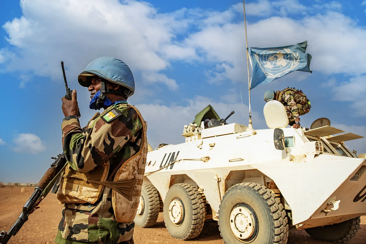 A Nigerian peacekeeper in uniform and a blue helmet talking into a radio holding a gun. In the background is a white tank flying the blue UN flag. They are both in the desert with blue sky with clouds behind them.