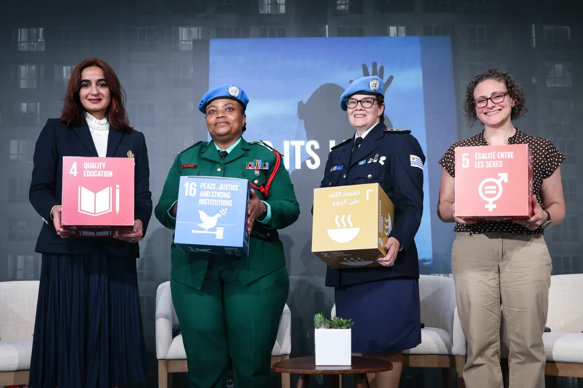 Four women peacekeepers stand side-by-side holding various SDGs