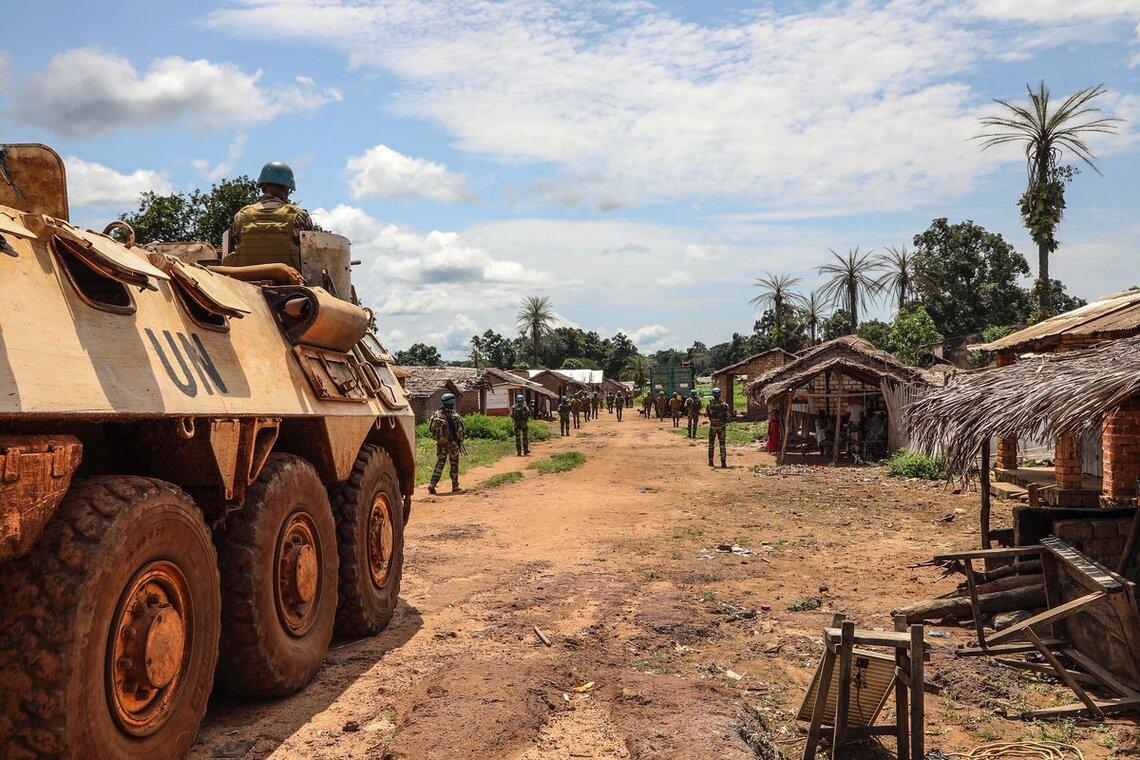 Un véhicule blindé des Nations Unies (ONU) est garé sur un chemin de terre dans un village rural. Le véhicule est de couleur beige, équipé de gros pneus et porte l'inscription « ONU » peinte sur le côté. Plusieurs soldats sont visibles, certains debout près du véhicule et d'autres plus loin sur la route.
